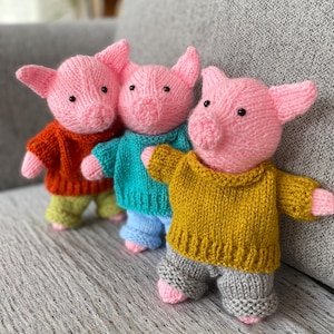 May include: Three knitted pig dolls, each with a pink body and black eyes. They wear different outfits: an orange sweater and green trousers, a teal sweater and blue trousers, and a yellow sweater with grey trousers.