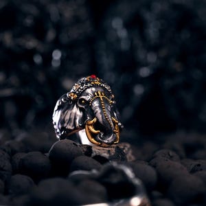 Może przedstawiać: Srebrny pierścień w kształcie głowy słonia, ze złotymi akcentami na trąbie i głowie. Mały czerwony klejnot znajduje się na szczycie głowy. Pierścień jest umieszczony na ciemnym, teksturowanym tle, tworząc uderzający kontrast.