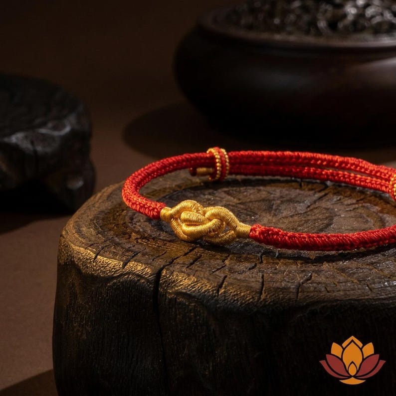 May include: A red and gold bracelet with a knot design. The bracelet is made of red cord with gold-colored accents. The knot is the focal point, and the bracelet is displayed on a wooden surface.