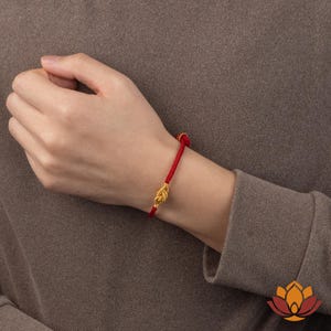 May include: A red cord bracelet with a gold-colored knot design. The bracelet is worn on a wrist, showcasing the vibrant red cord and the intricate knot detail. The background is a solid, neutral color.