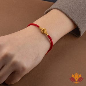 May include: A red cord bracelet with a gold-colored knot design. The bracelet is worn on a wrist, showcasing the contrast between the vibrant red cord and the metallic knot. The bracelet is a simple yet elegant piece of jewelry.