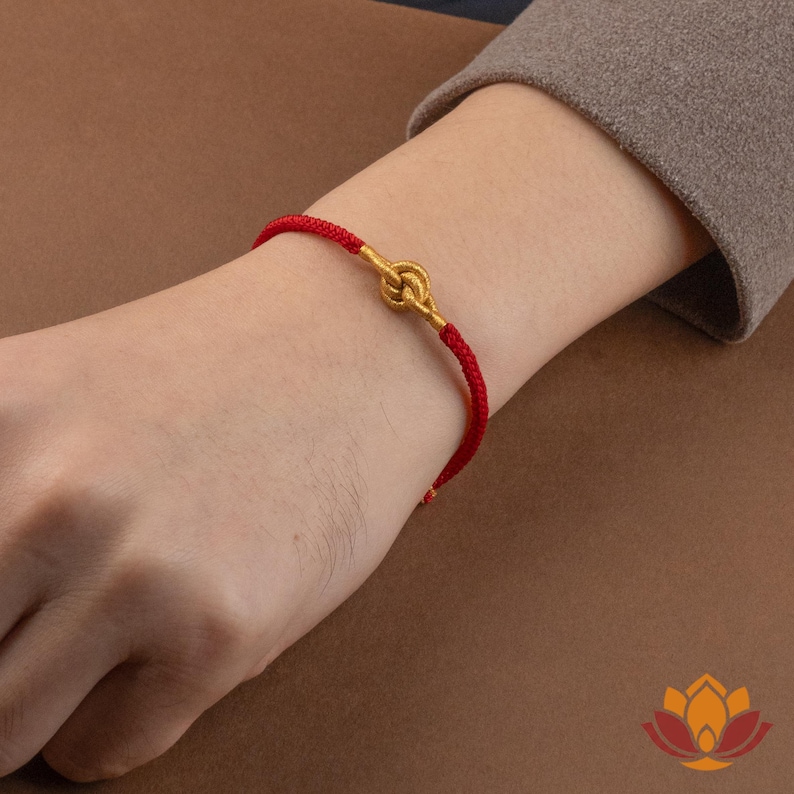 May include: A red cord bracelet with a gold-colored knot design. The bracelet is worn on a wrist, showcasing the contrast between the vibrant red cord and the metallic knot. The bracelet is a simple yet elegant piece of jewelry.