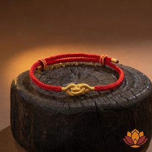 May include: A red and gold bracelet with a knot design. The bracelet features a red cord with gold accents and a central gold knot. The bracelet is displayed on a dark wooden surface.