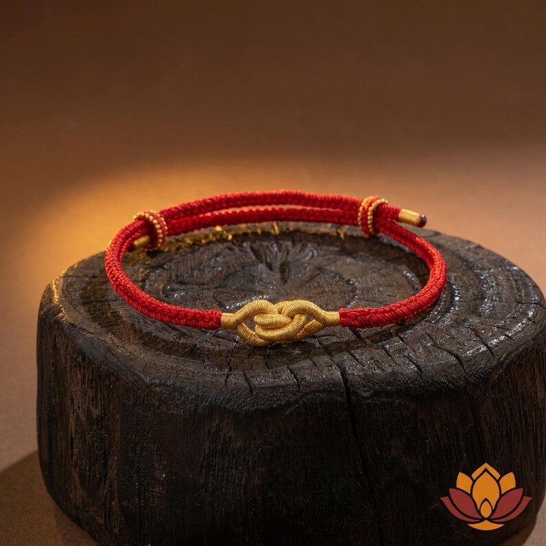 May include: A red and gold bracelet with a knot design. The bracelet features a red cord with gold accents and a central gold knot. The bracelet is displayed on a dark wooden surface.