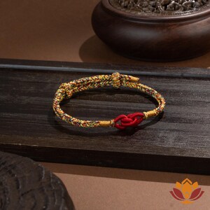 May include: A braided bracelet with a red knot closure. The bracelet features a mix of yellow, green, and red threads. The background includes a dark wooden surface and a decorative wooden bowl.