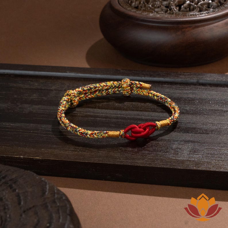 May include: A braided bracelet with a red knot closure. The bracelet features a mix of yellow, green, and red threads. The background includes a dark wooden surface and a decorative wooden bowl.