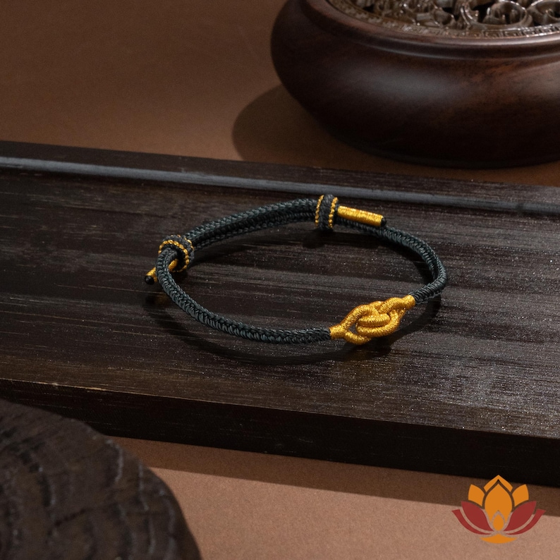 May include: A black and gold bracelet with a woven cord design. The bracelet features gold-colored accents at the clasp and a decorative knot in the center. The bracelet is displayed on a dark wooden surface.