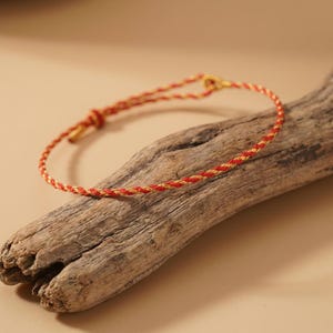 May include: A red and gold braided bracelet with a gold clasp. The bracelet is displayed on a piece of weathered wood against a neutral background. The bracelet's design features a twisted rope pattern.