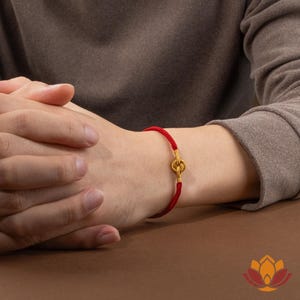 May include: A red bracelet with a gold-colored clasp is worn on a wrist. The bracelet is made of a woven red cord. The clasp is a decorative gold-colored design. The person is wearing a brown long-sleeved shirt.