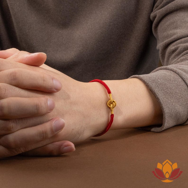 May include: A red bracelet with a gold-colored clasp is worn on a wrist. The bracelet is made of a woven red cord. The clasp is a decorative gold-colored design. The person is wearing a brown long-sleeved shirt.