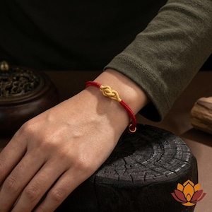 May include: Red and gold bracelet featuring a knot design. The bracelet has a red cord with a gold-colored knot in the center. The bracelet is worn on a wrist, with a dark background.