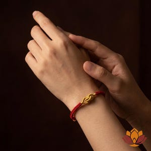 May include: A red cord bracelet with a gold-colored knot design. The bracelet is worn on a wrist, with hands in the background. The bracelet's knot is the focal point, showcasing a simple yet elegant design.