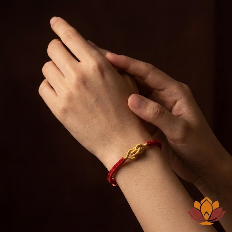 May include: A red cord bracelet with a gold-colored knot design. The bracelet is worn on a wrist, with hands in the background. The bracelet's knot is the focal point, showcasing a simple yet elegant design.