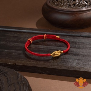 May include: A red bracelet with a gold knot clasp and gold accents. The bracelet is made of a woven red material and is displayed on a dark wooden surface. The bracelet is a piece of jewelry.