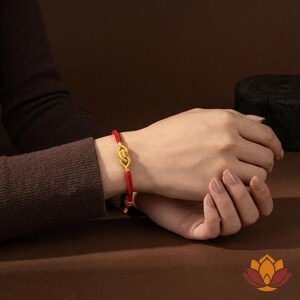 May include: A gold-colored bracelet with a knot design is displayed on a wrist. The bracelet is attached to a red cord. The background is a solid brown color. The bracelet is a piece of jewelry.