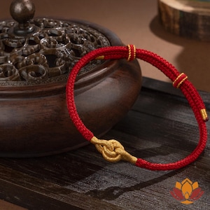 Puede incluir: Una pulsera roja con detalles dorados. La pulsera presenta un cordón rojo tejido con nudos dorados y pequeñas cuentas doradas. Se muestra sobre una superficie de madera, con un recipiente decorativo de madera en el fondo.
