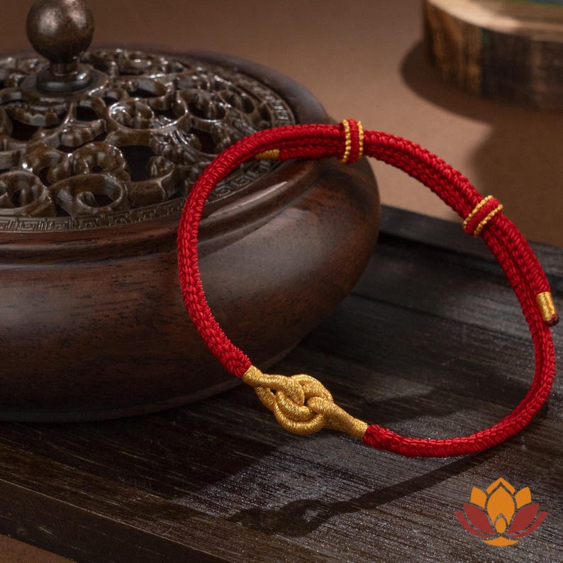 May include: A red bracelet with gold accents. The bracelet features a woven red cord with gold-colored knots and small gold beads. The bracelet is displayed on a wooden surface, with a decorative wooden container in the background.
