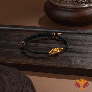 May include: Black braided bracelet with gold-colored accents. The bracelet features a knot design in the center and gold-colored details at the ends. The bracelet is displayed on a dark wooden surface.