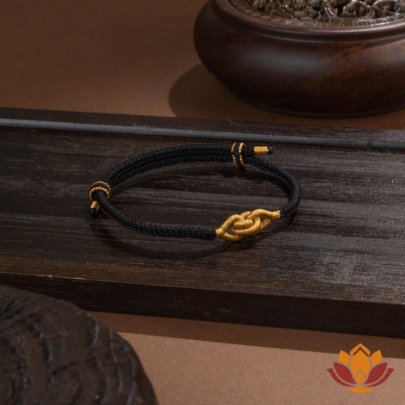 May include: Black braided bracelet with gold-colored accents. The bracelet features a knot design in the center and gold-colored details at the ends. The bracelet is displayed on a dark wooden surface.