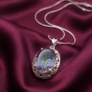 May include: A silver pendant necklace featuring a large, oval-cut mystic topaz gemstone with iridescent rainbow hues. The stone is set in a decorative, openwork silver frame with intricate scrollwork details, suspended from a thin silver chain.