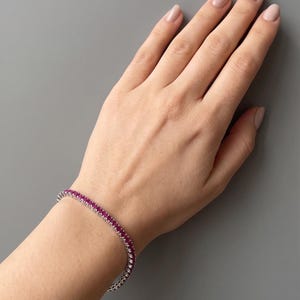 May include: A tennis bracelet featuring a continuous row of small, vibrant pinkish-red gemstones set in a polished silver-toned metal band. The bracelet encircles the wrist, showcasing a uniform design with each stone securely held in a metal setting.