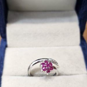 Op de afbeelding: Een zilverkleurige ring met een bloemvormig cluster van zes ronde, dieproze robijnen in het midden. De ringband is glad en gepolijst, met een klein gedeelte bij de bloemvormige zetting dat is versierd met diverse kleine, heldere steentjes.
