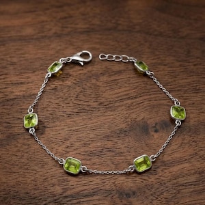 May include: A silver bracelet featuring a series of square-cut, light green gemstones. The bracelet is composed of a delicate chain with the gemstones evenly spaced along its length. The clasp is visible at one end. The bracelet is displayed on a dark wood surface.