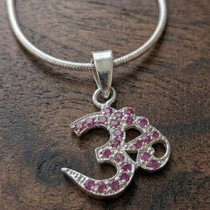 May include: A silver-colored pendant necklace featuring the Om symbol, encrusted with small, pink gemstones. The pendant hangs from a thin, silver chain. The background is a dark, wooden surface.
