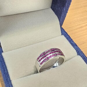 May include: A silver-toned ring featuring three parallel rows of small, square-cut, vibrant pink gemstones set into the band. The ring is displayed inside a blue velvet-lined jewellery box with a light-coloured interior cushion.