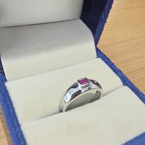 May include: A silver-toned ring featuring a square-cut red gemstone set in the centre, flanked by two small, round, clear stones on either side of the band. The ring is displayed inside a dark blue velvet-lined jewellery box with a light interior.