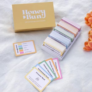 May include: A yellow box with the text "Honey Bun" and a deck of colourful cards. The cards are in a white box, and some are fanned out. Orange tulips are on the right side of the image. The cards have text.
