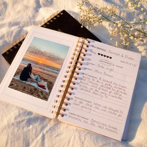 Könnte beinhalten: Ein offenes Spiralbuch mit einem Foto eines Sonnenuntergangs am Strand und handschriftlichen Notizen. Das goldfarbene Notizbuch liegt auf einem weißen Tuch, im Hintergrund ein schwarzes Notizbuch. Ein Zweig mit weißen Blüten befindet sich oben rechts.