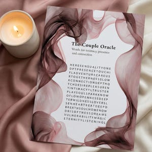 Könnte beinhalten: Eine brennende Kerze in einem weißen Keramikhalter steht neben einer Karte mit dem Titel "The Couple Oracle". Die Karte zeigt ein wirbelndes Design in Maroon und Rosa mit Wörtern über Intimität und Verbindung. Die Karte liegt auf einer rosa Satin-Oberfläche.