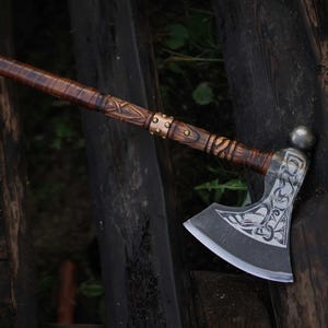 May include: A handcrafted axe with a dark wooden handle and a silver-colored blade. The handle features intricate carvings and leather wrapping. The axe head has decorative engravings. The axe is leaning against a weathered wooden surface.
