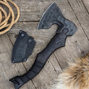 Puede incluir: Un hacha negra con cabeza y mango texturizados, sobre una superficie de madera. La cabeza del hacha tiene un patrón hexagonal único. Una funda de cuero negro está junto al hacha. Una bobina de cuerda y piel también están en la imagen.