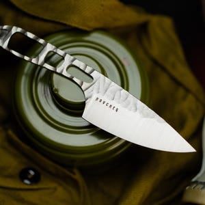 May include: A small, silver knife with a textured blade and a patterned handle. The word "NOUCHER" is etched on the blade. The knife rests on a green object, possibly a gas mask filter, with a khaki-coloured background.