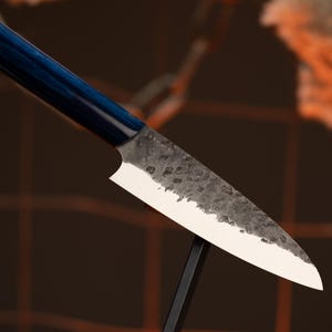 Puede incluir: Un cuchillo de cocina de estilo japonés con un mango azul oscuro y una hoja texturizada de color plateado. El cuchillo se exhibe sobre un soporte negro contra un fondo borroso, mostrando su filo y artesanía.