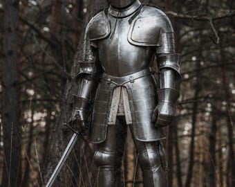 May include: A full suit of medieval armor, including a helmet, chest plate, and leg guards, stands in a forest setting. The silver-colored armor reflects the light, and a sword is held at the side. The background features tall trees and dry grass.