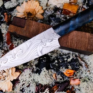 Puede incluir: Un cuchillo artesanal con una hoja estampada y un mango azul y negro descansa sobre un bloque de madera. La hoja presenta un diseño en forma de ola. El cuchillo está rodeado de flores secas y musgo.