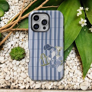 Pode incluir: Uma capa de telemóvel às riscas azuis e brancas com um estampado gráfico de um boné, um saco, um biquíni, um copo de martini e um jarro com flores. A capa tem um recorte para um sistema de câmara tripla no canto superior esquerdo.