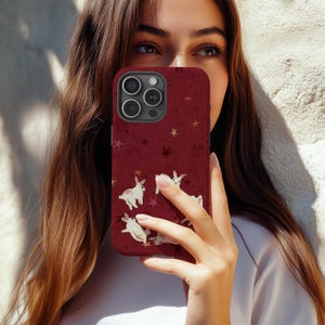 May include: A burgundy phone case featuring a pattern of white rabbits and gold stars. The case has a textured appearance and a dark camera lens area. The design is set against a deep red background.