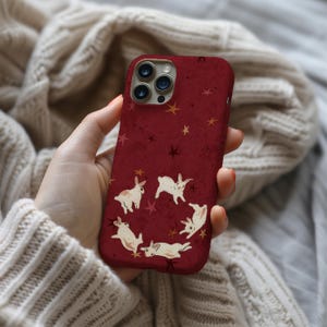 May include: A burgundy phone case featuring a whimsical design of white rabbits arranged in a circle, with scattered gold and dark red stars. The case has a glossy finish and is designed for a smartphone.