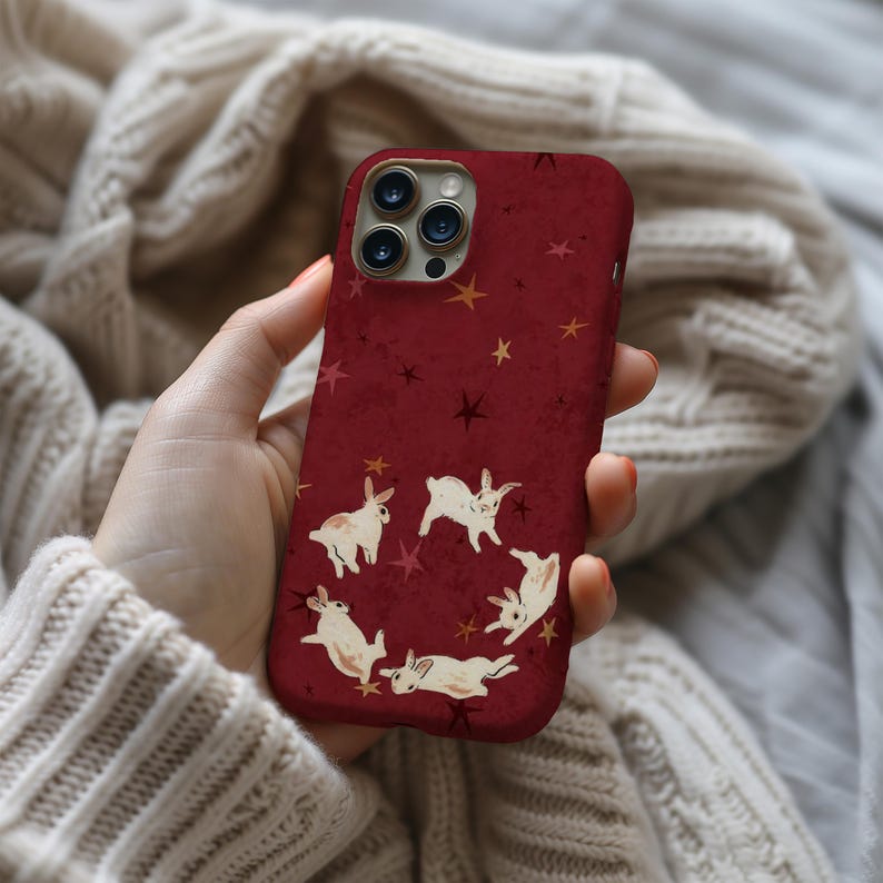 May include: A burgundy phone case featuring a whimsical design of white rabbits arranged in a circle, with scattered gold and dark red stars. The case has a glossy finish and is designed for a smartphone.