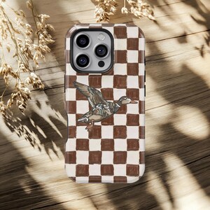 May include: A phone case with a brown and white chequered design, showcasing a detailed illustration of a duck in flight. The case has a clear border and a camera cut-out.
