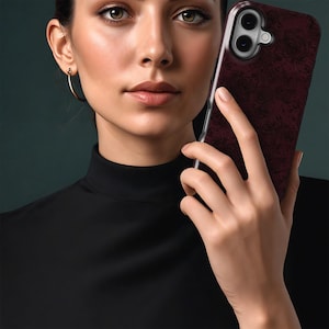 May include: A burgundy phone case with a floral design is held in front of a person. The case has a clear border and a camera cutout. The person wears a black turtleneck and gold hoop earrings.