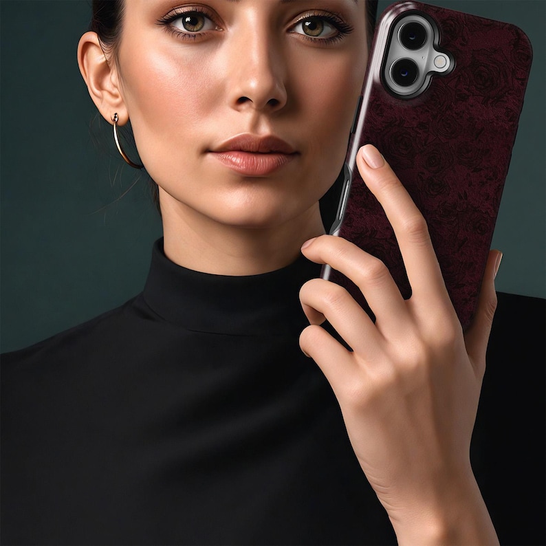 May include: A burgundy phone case with a floral design is held in front of a person. The case has a clear border and a camera cutout. The person wears a black turtleneck and gold hoop earrings.