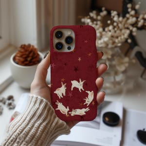 May include: A burgundy phone case featuring a pattern of white rabbits and gold stars. The case has a textured appearance and a camera cutout. The design is repeated on the back of the phone case.