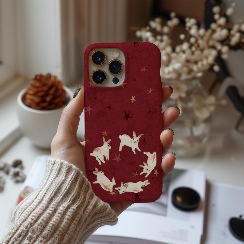 May include: A burgundy phone case featuring a pattern of white rabbits and gold stars. The case has a textured appearance and a camera cutout. The design is repeated on the back of the phone case.