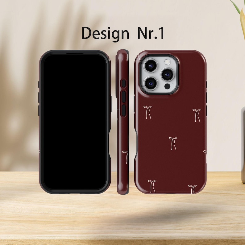May include: Three phone cases are displayed. The front of the phone case is black, the side is maroon, and the back is maroon with a pattern of small, light-coloured bows. The text "Design Nr.1" is at the top.