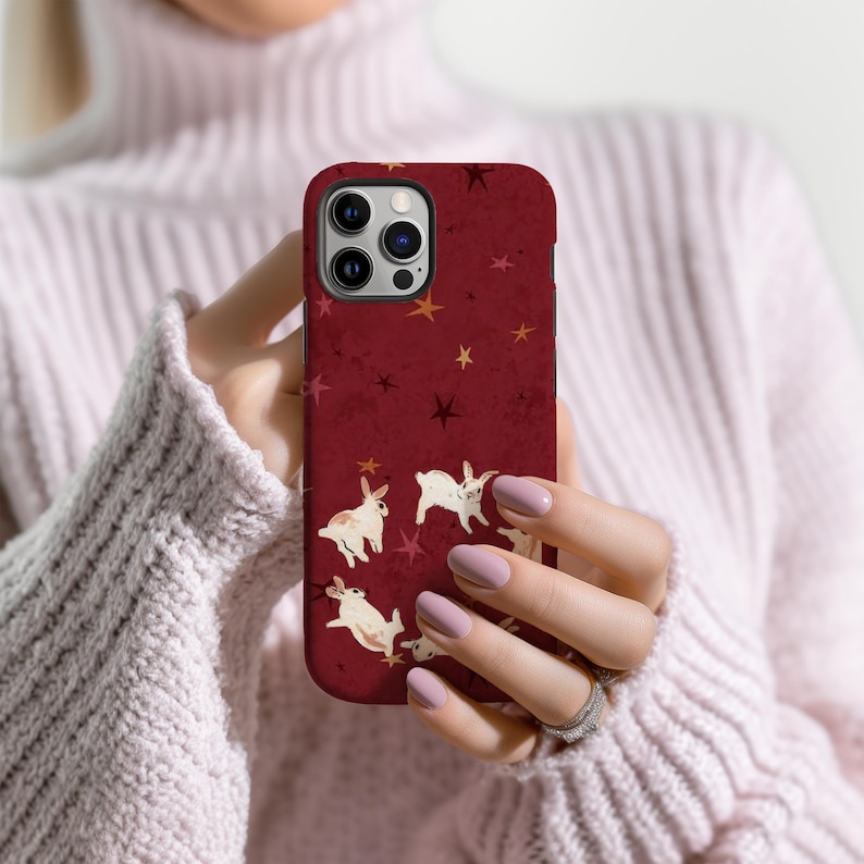 May include: A burgundy phone case featuring a pattern of white rabbits and stars. The case has a textured appearance and a dark border. The design is repeated across the surface of the case.